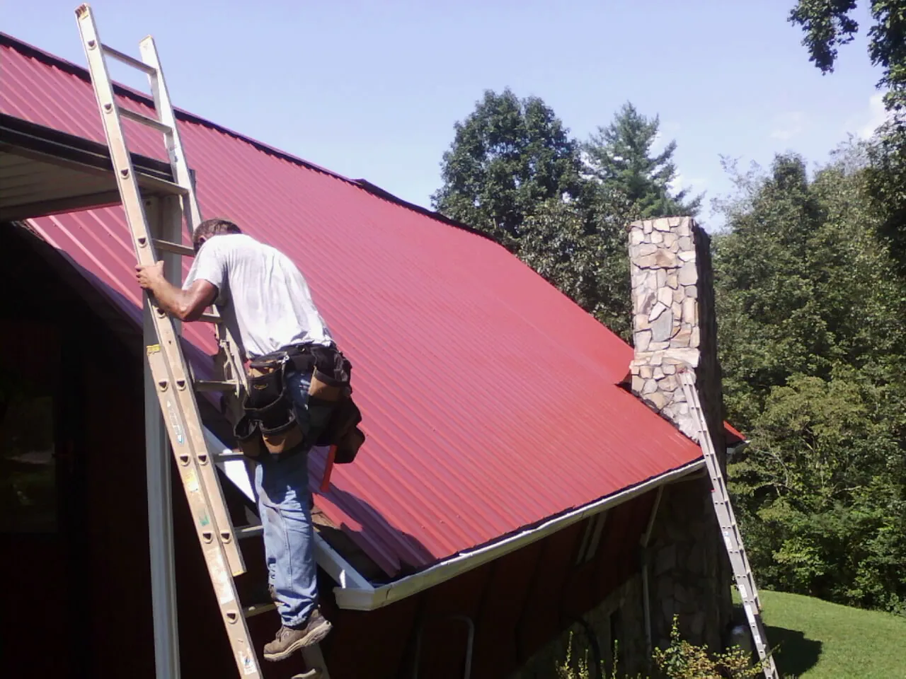 Roofer on a ladder beside a red metal roof and stone chimney — Soffit & Fascia Repair by Roofing Brothers in Red Springs